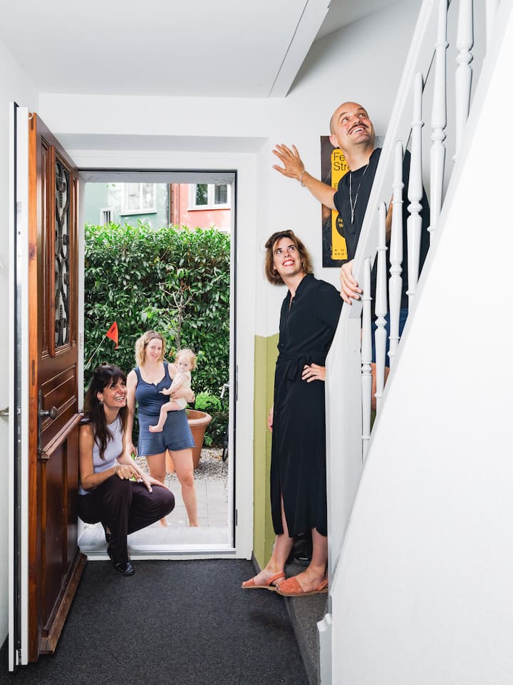 Vier Leute einer WG stehen in einem Treppenhaus - Steigende Immobilienpreise und strenge Bankenregeln machen den Hauskauf selbst für Gutverdienende fast unmöglich. Doch innovative Wohnprojekte zeigen, wie gemeinschaftliches Leben bezahlbaren Wohnraum schafft