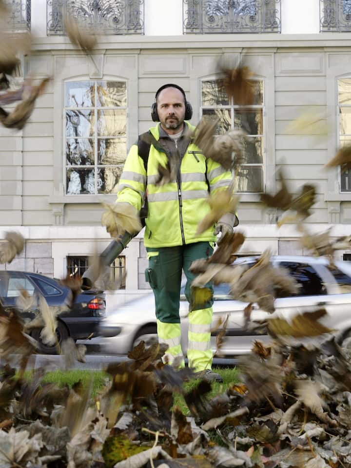 Ein Arbeiter wirbelt mit dem Laubbläser Herbstlaub auf einer Wiese auf.