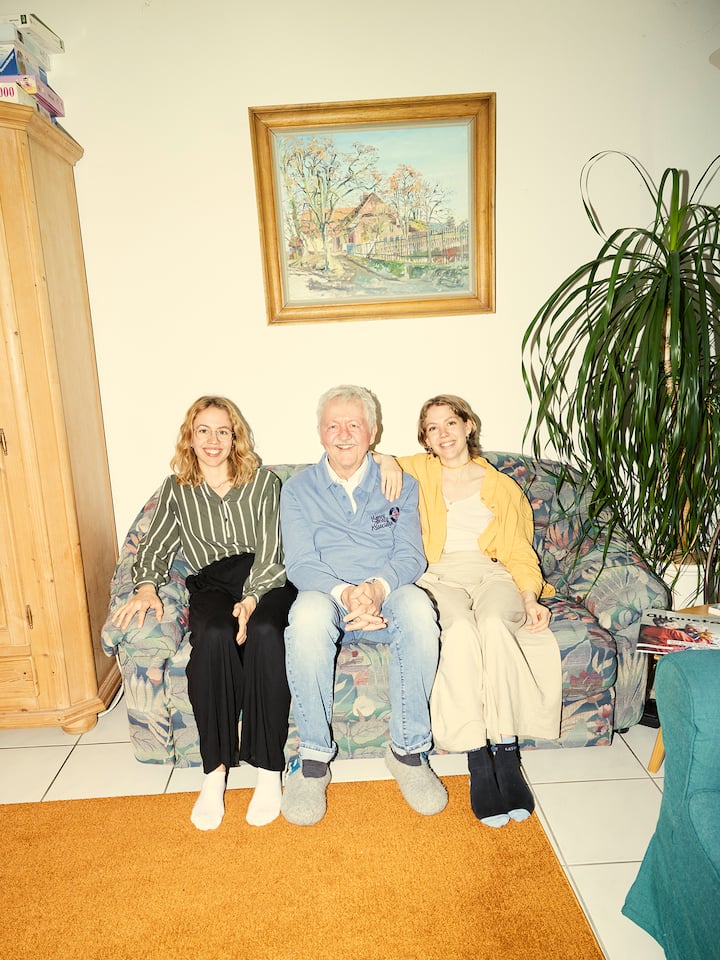 Myrjam, Johanna und ihr Vater sitzen lächelnd auf dem Sofa - Myrjam und Johanna waren Teenager, als ihr Vater an Demenz erkrankte. Heute betreuen sie ihn gemeinsam mit ihrer Mutter zuhause. Wie schaffen sie das, ohne dabei das Erwachsenwerden zu verpassen?