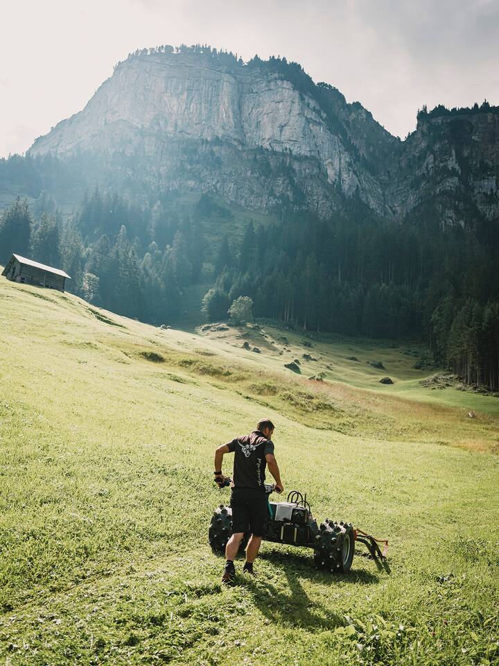 Ein Mann schiebt ein Landwirtschaftsgerät auf einer grünen Bergwiese, im Hintergrund hohe Berge.