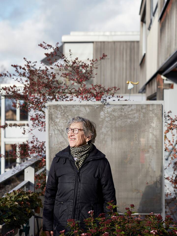 Eine ältere Frau mit Brille und kurzen grauen Haaren steht auf einem Balkon und lächelt in die Sonne.