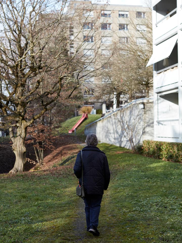 Man sieht eine ältere rüstige Frau von hinten, wie sie in einer Wohnsiedlung zwischen Baum und Gebäude einen Weg durch den Rasen hochgeht, im Hintergrund ein Kinderspielplatz mit roter Rutschbahn.