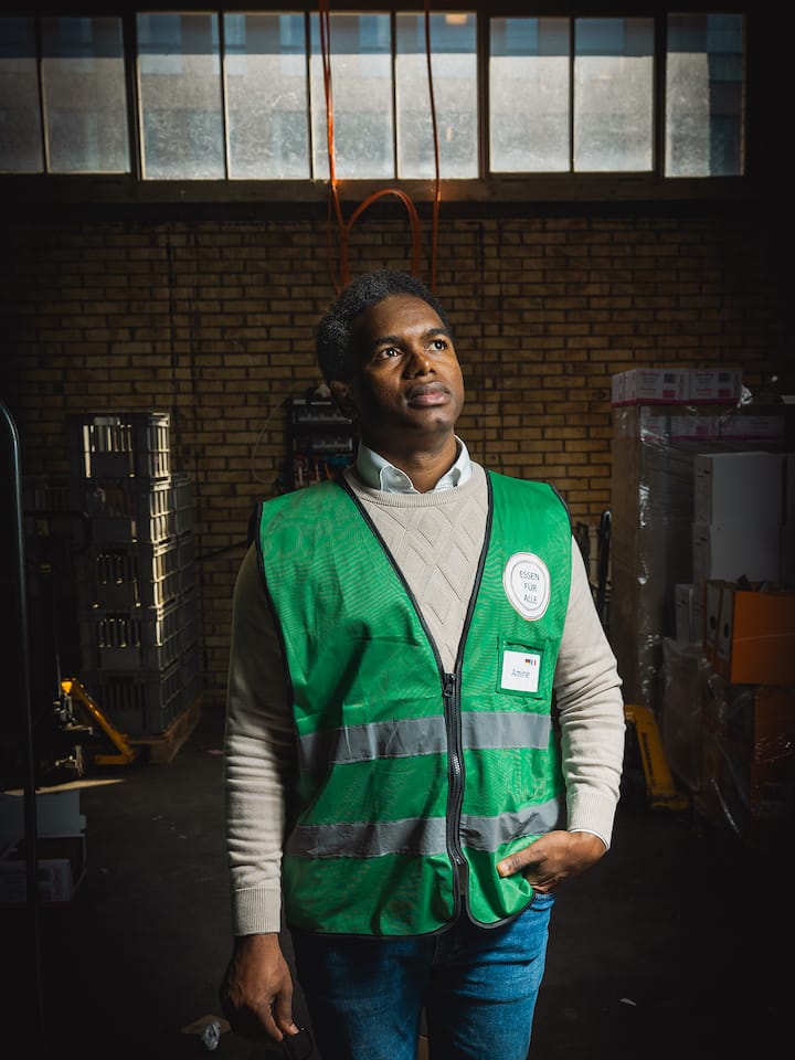 Amine Diare Conde steht in einer Lagerhalle, er blickt seitlich nach oben und trägt eine grüne Arbeitsweste.