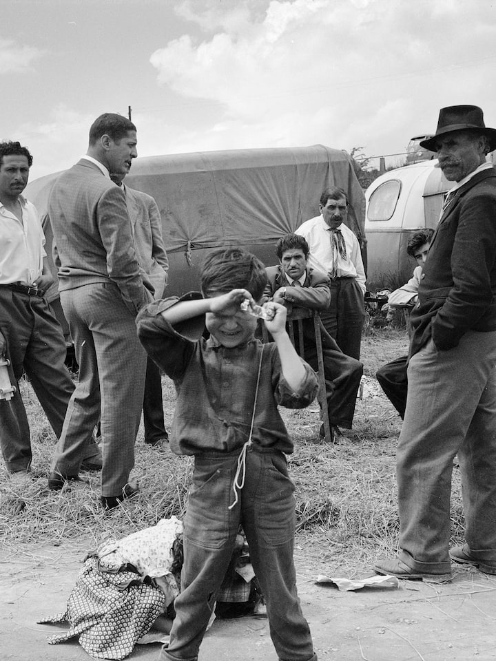 Am Stadtrand von Zürich haben Fahrende einen Standplatz neben den Bahngeleisen, aufgenommen im Jahr 1958. Männer stehen zum Gespräch im Kreis. Ein Junge im Vordergrund verdeckt mit seinen Händen sein Gesicht.