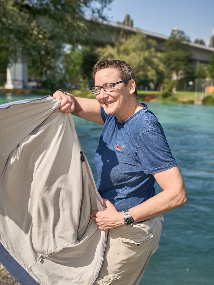 Chantal Britt steht an der Aare, lächelt in die Kamera und schwingt ihre Jacke zum Anziehen nach hinten.