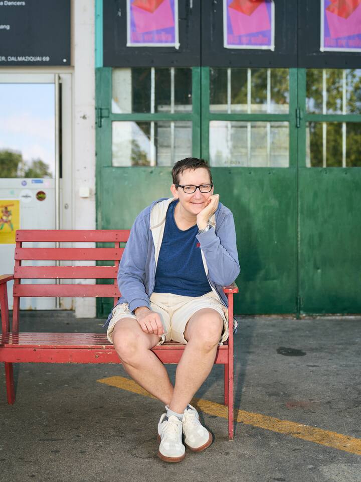Chantal Britt sitzt auf einer roten Bank vor einer grünen Türe, sie stützt sich mit dem Ellbogen auf der Lehne auf, ihre Hand stützt das Kinn, sie trägt Brille und lächelt direkt in die Kamera.