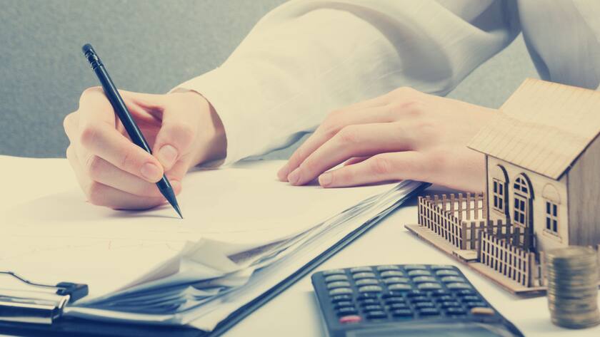 Was gilt bei den Steuern? Es ist eine Hand zu sehen, die mit einem Kugelschreiber ein Formular ausfüllt. Daneben liegt noch ein Taschenrechner und ein kleines Modell eines Einfamilienhauses.