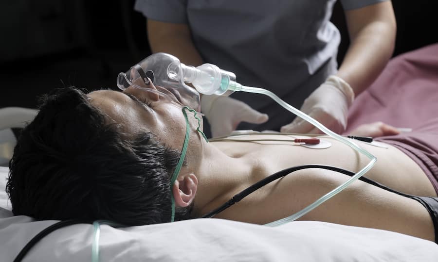 Ein Mann mit einer Sauerstoffmaske liegt in einem Bett und wird von einer Person mit Handschuhen betreut.