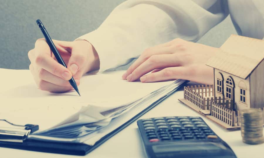 Es ist eine Hand zu sehen, die mit einem Kugelschreiber ein Formular ausfüllt. Daneben liegt noch ein Taschenrechner und ein kleines Modell eines Einfamilienhauses.
