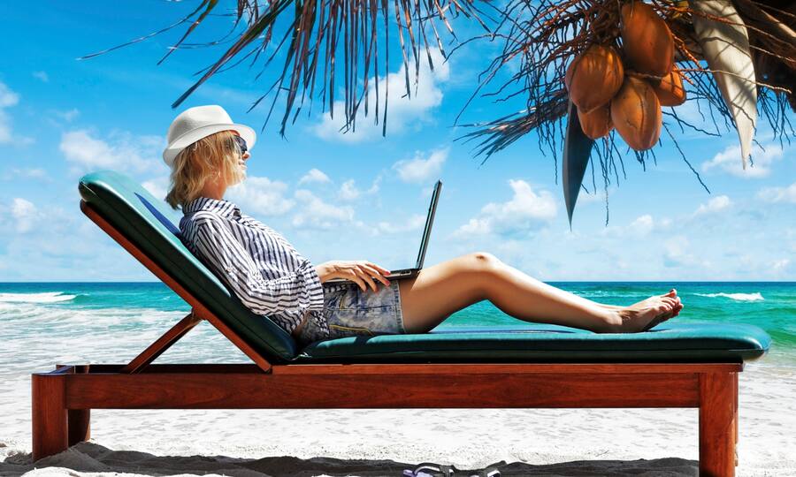 Frau liegt mit Laptop in der Hand auf einem Liegestuhl am Strand unter Palmen