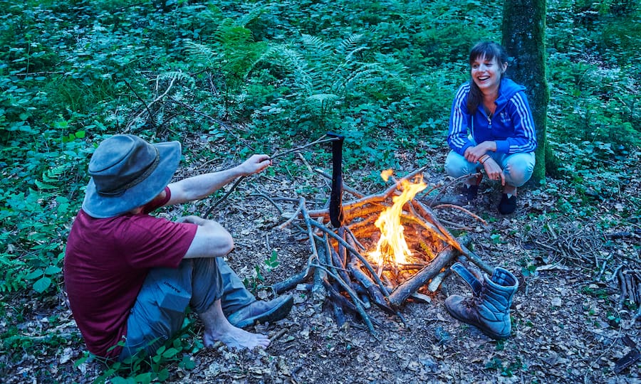 Zwei Waldcamper entzünden ein Feuer, dessen Holz zu einer Art Vogelnest zusammengelegt wurde.