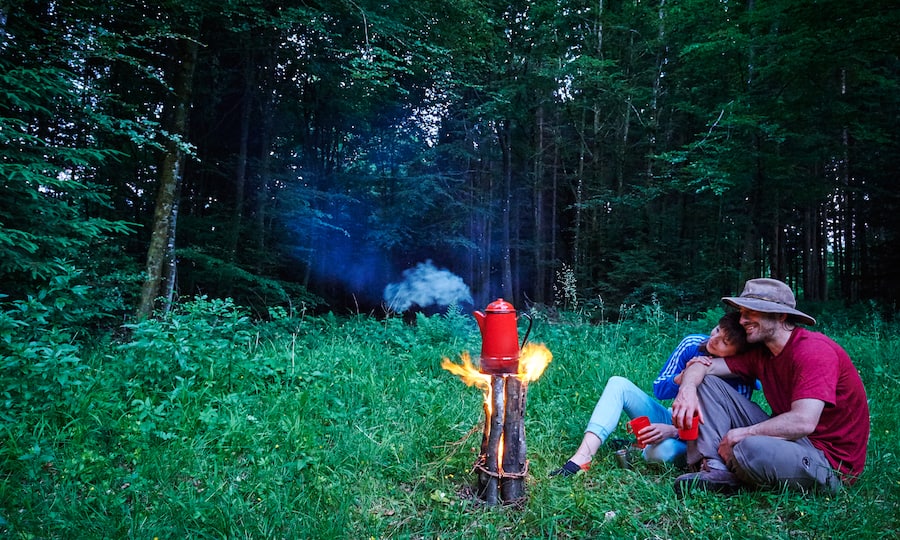 Zwei Waldcamper sitzen auf einem Boden mit hohem Gras und schauen dabei zu, wie eine heisse Flüssigkeit in einer Kanne über einer Feuerstelle zu kochen beginnt.