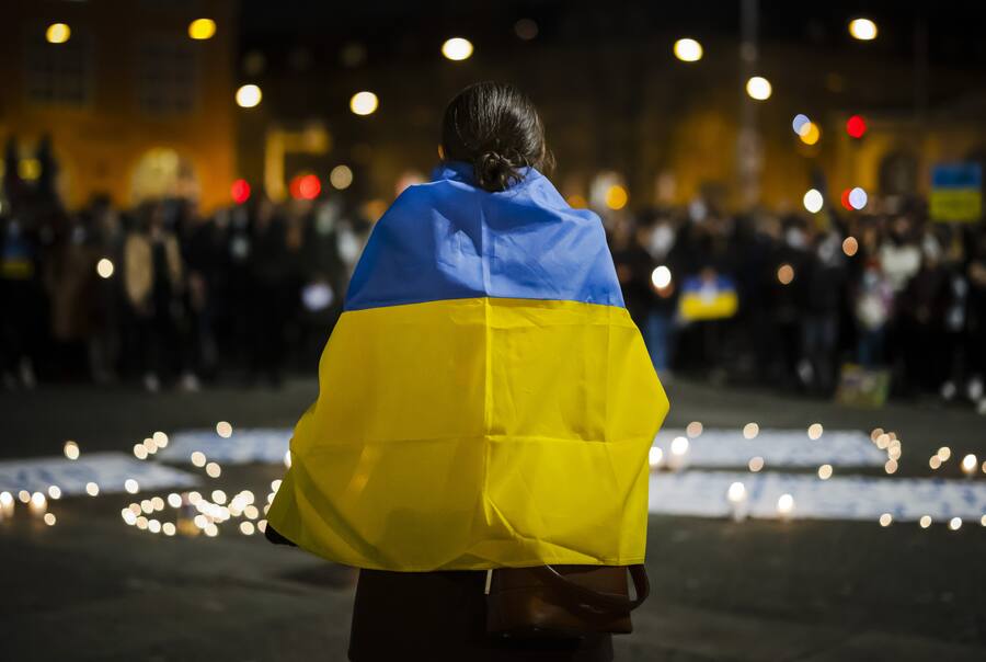 Eine Frau trägt eine ukrainische Flagge und spricht vor einem Meer aus Kerzen und den versammelten Demonstranten ihre Solidarität mit den Ukrainerinnen und Ukrainern aus, die vom russischen Angriffskrieg betroffen sind.