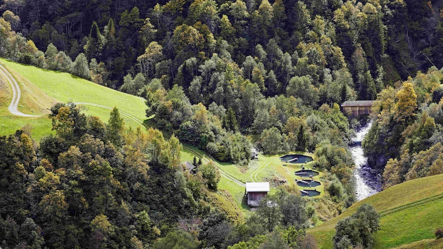 Die vier Zuchtbecken der Fischzuchtanlage von Curdin und Sarah Capeder mit dem Fluss Glogn von der Kapelle Sogn Roc in Lumbrein aus gesehen. (Das Wasser für die Anlage stammt nicht aus dem Fluss der im Bild sichtbar ist)