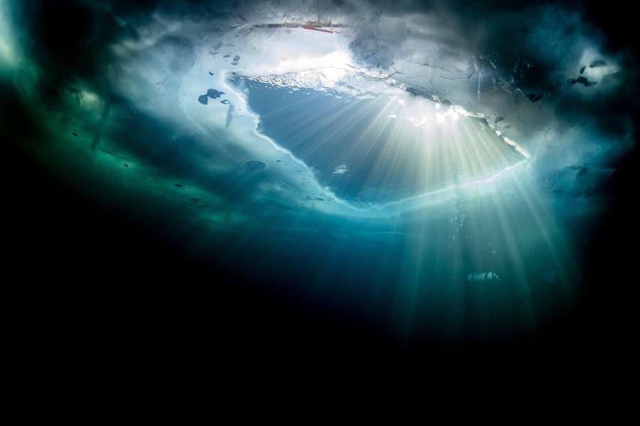 Sonnenlicht scheint durch ein Loch in der Eisdecke in den kalten See.  Diesem Lichtstrahl kann sich niemand entziehen: Eisdecke des Silsersees von unten, 2022. Ein Sprung unters Eis löst bei vielen Panik aus. Doch wer die Angst besiegt, wird mit einem überirdischen Erlebnis belohnt.