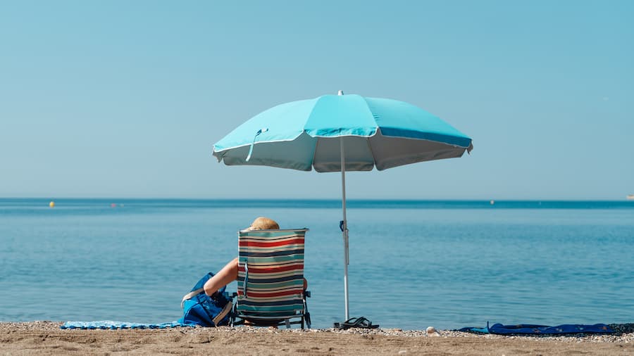 Eine Frau sitzt in einem Liegestuhl unter einem blauen Sonnenschirm am Strand, im Hintergrund das blaue Meer.