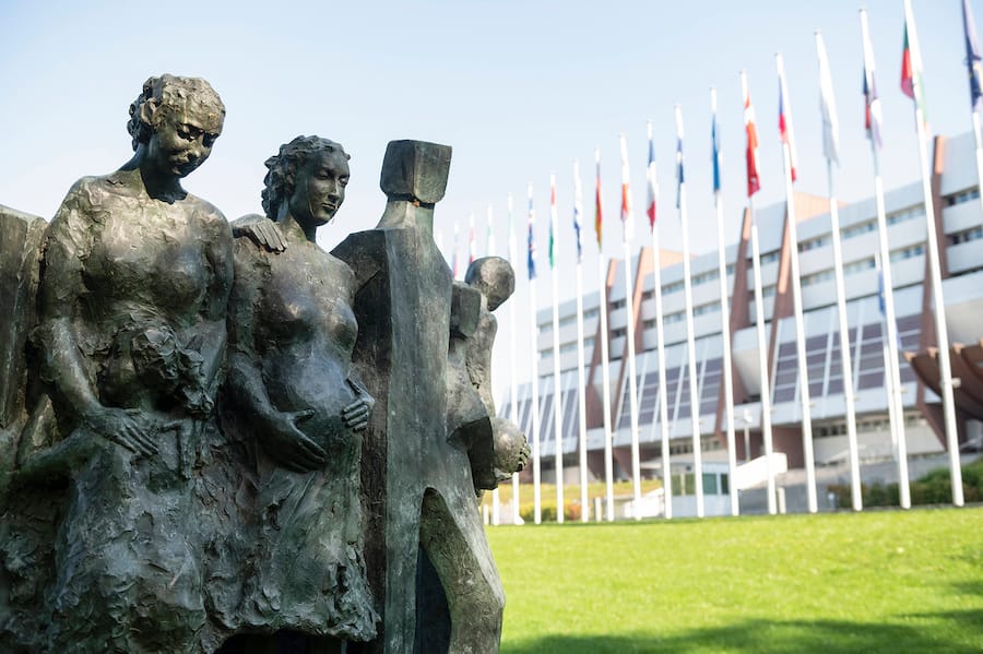 Eine Statue mit Menschenfiguren vor einer Reihe von Flaggen vor dem Gebäude des Europarates in Strassburg.