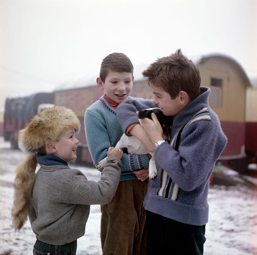 Drei Kinder in Winterpullovern halten einen Welpen in der Hand, sie stehen vor einem Wohnwagen, ein Kind trägt eine Fellmütze.