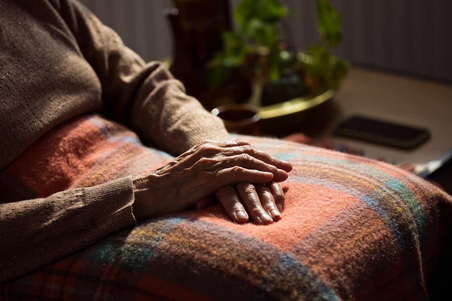 Senior woman sitting in armchair at home with legs covered by blanket. Close up of hands, unrecognizable person.